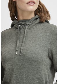 Sudadera verde hecha de una tela suave, con capucha ajustable y cordones con puntas de plata, de corte relajado. La superficie tiene una textura lisa.