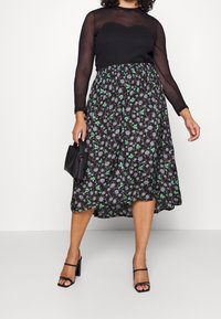 Abito nero con una blusa trasparente a maniche lunghe e una gonna con motivo floreale caratterizzata da tonalità di verde e viola. Borsa nera e sandali con cinturini.