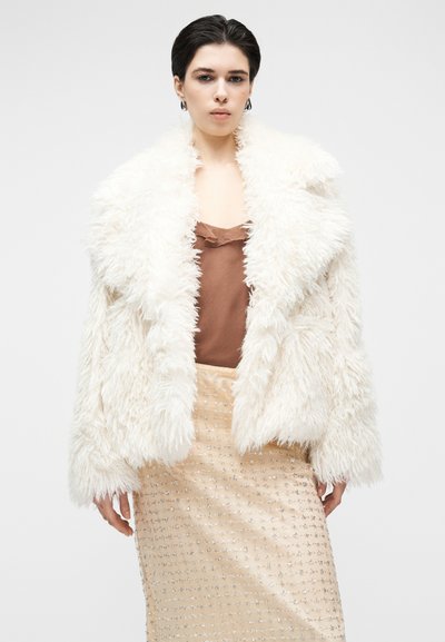 Veste en fausse fourrure blanche avec col oversize, associée à un haut marron et une jupe beige ornée de sequins argentés.