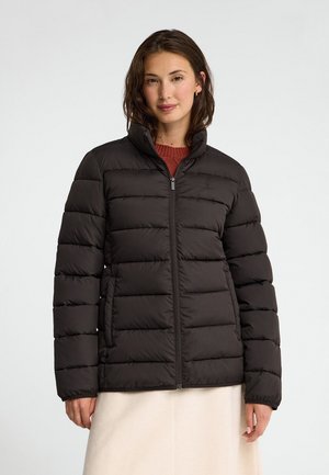 Donna che indossa una giacca imbottita nera con cerniera, in piedi contro uno sfondo chiaro uniforme, con una gonna beige e un maglione color ruggine visibili.