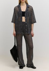 Chiffon tweedelige outfit met een marineblauw en crèmekleurig zigzagpatroon, oversized shirt met korte mouwen, aansluitende broek en een zwarte totebag.