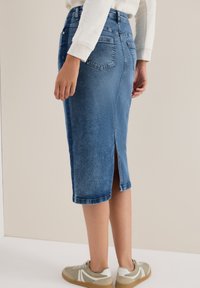 Denim kokerrok in donkerblauw, midi-lengte met een split aan de achterkant, vijf zakken en een vervaagde wassing, gecombineerd met beige sneakers.