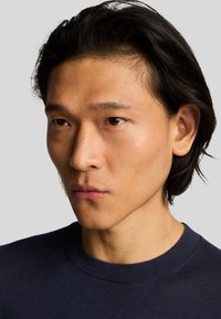 Primo piano di un volto maschile con capelli scuri e lisci, che indossa un maglione girocollo blu scuro. Texture della pelle liscia e espressione neutra.