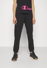 Pantalones de chándal negros con puños elásticos, combinados con una camiseta sin mangas negra que presenta un gran logo rosa. Llevado con zapatillas deportivas negras.