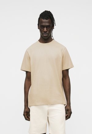 Homme à la peau foncée et aux cheveux tressés, portant un t-shirt beige à manches courtes et un short blanc, debout devant un fond clair uni.