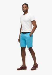 Pantalones cortos de algodón azul con bolsillos laterales, combinados con una camiseta blanca de cuello redondo y zapatos de barco de cuero marrón. Cinturón y reloj marrones como acento.