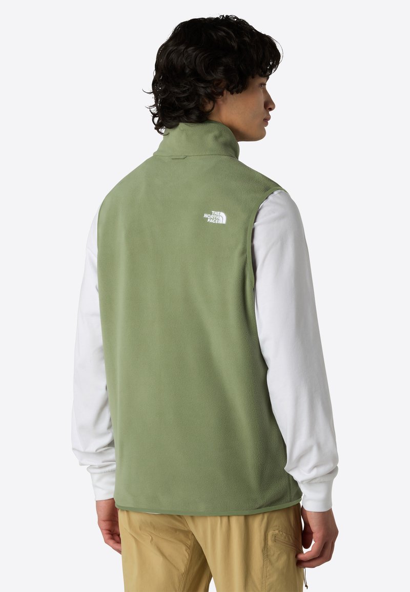 Fleece-Weste in einem gedeckten Grün, mit hohem Kragen und weißem Logo auf dem oberen Rücken. Getragen über einem langeärmeligen weißen Shirt.