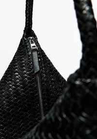 Schwarze gewebte Handtasche mit strukturiertem Design und glänzendem Metallreißverschluss. Verfügt über einen robusten Griff und eine zulaufende Form.