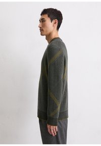 Homme en pull à motif vert foncé et pantalon gris se tenant de profil contre un mur blanc uni.
