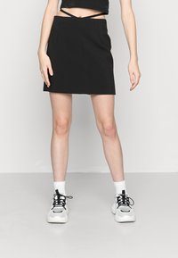 Mini jupe noire avec deux bretelles découpées à l'avant, tissu lisse, silhouette ajustée, associée à des chaussettes blanches et des baskets noires épaisses.
