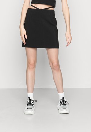Mini jupe noire avec deux bretelles découpées à l'avant, tissu lisse, silhouette ajustée, associée à des chaussettes blanches et des baskets noires épaisses.