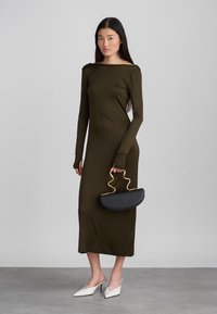 Olívazöld hosszú ujjú ruha csónaknyakkivágással, fekete ívelt kézitáskával, arany hullámos fogantyúval, valamint fehér hegyes orrú slip-on cipőkkel bemutatva.