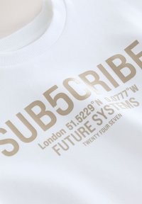 Biela mikina s reflexným béžovým textom s nápisom "SUB5CRIBE", súradnicami Londýna, "FUTURE SYSTEMS" a "TWENTY FOUR SEVEN".