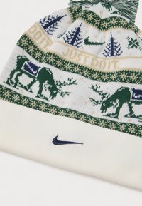 Pleteni beli zimski klobuk z zelenimi in modrimi motivi jelena in dreves, besedilom "SAMA TO NAREDI" ter modrimi logotipi Nike swoosh, na vrhu s pom-pomom.