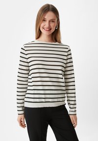 Sweter z długim rękawem w kremowym kolorze w ciemnozielone paski. Miękka tkanina, okrągły dekolt i płaski dół. Noszony z czarnymi spodniami.