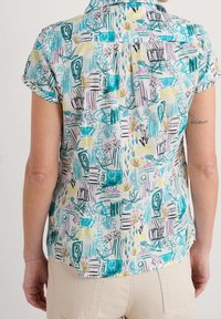 Chemise à manches courtes avec un motif floral abstrait coloré en tons de bleu sarcelle, violet et jaune sur un fond clair ; tissu léger.