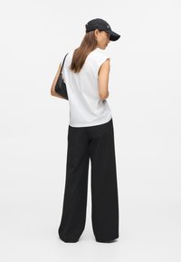 Γυναίκα φορώντας μαύρο καπέλο, λευκό αμάνικο τοπ και φαρδιά μαύρα παντελόνια, κρατώντας μια μαύρη τσάντα χεριού, στέκεται μπροστά σε λευκό φόντο.