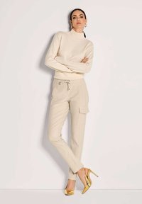 Vrouw in een beige coltrui en bijpassende cargobroek staat met gekruiste armen, gekleed in glanzende gouden high heels.