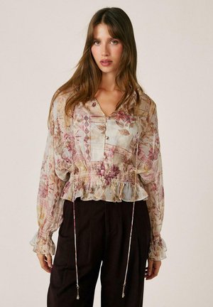 Mujer con el pelo largo y castaño que lleva una blusa floral transparente con puños con volantes y pantalones negros de cintura alta sobre un fondo liso.