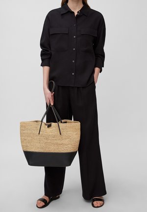 Camicia nera a bottoni con due tasche sul petto, abbinata a pantaloni neri larghi. Il modello tiene una borsa a spalla intrecciata di paglia color beige con fondo nero.