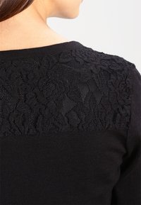 Svart stickad tröja med en floralkönsnätbakpanel, med rund halsringning och slät textur, som erbjuder en kontrasterande genomskinlig design.