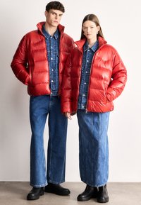 Κόκκινα μπουφάν puffer με γυαλιστερό φινίρισμα, συνδυασμένα με μπλε πουκάμισα από τζιν. Τα φιγούρες φορούν παντελόνια με φαρδιά πόδια και μαύρα μποτάκια με πλατφόρμα.