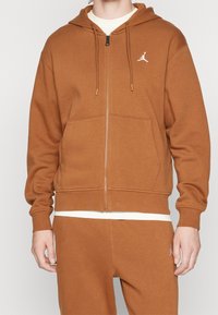 Felpa con zip marrone, dotata di cappuccio, tasche frontali e un piccolo logo bianco sul petto. Tessuto morbido con vestibilità rilassata, polsini e orlo a coste.