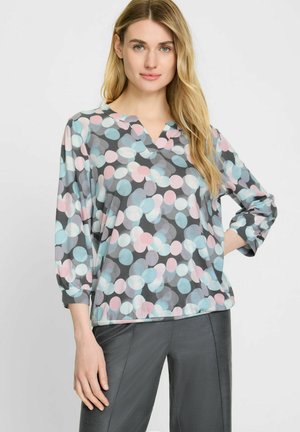 Mehrfarbige Bluse mit V-Ausschnitt, die ein Muster aus rosa, hellblauen und grauen undurchsichtigen Kreisen auf einem dunkelgrauen Hintergrund aufweist.