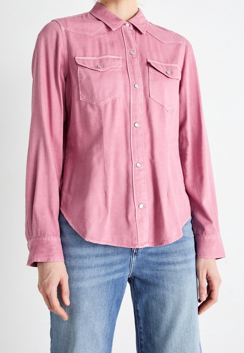 Chemise à manches longues, boutonnée, en tissu rose clair. Dispose de deux poches poitrine avec rabat et fermetures par pressions, ourlet arrondi et col simple.