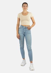 Beige kurz geschnittenes T-Shirt, hellblaue hochgeschnittene Jeans und weiße Sneakers. Das Model steht mit einer Hand in der Tasche vor einem neutralen Hintergrund.