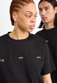 Camiseta de algodón negra con texto verde neón, cuello redondo y mangas cortas, acompañada de un fondo con otra persona vestida con una camiseta negra.