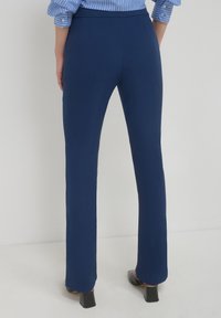 Person in navyblauen, geraden Hosen, schwarzen High Heels und einem blau gestreiften Hemd mit hochgekrempelten Ärmeln.