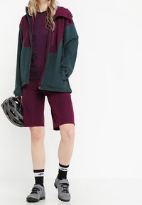 Sportoutfit mit einer bordeauxroten und dunkelgrünen Zip-Jacke, passenden bordeauxroten Shorts, grauen Schuhen und einem Fahrradhelm.