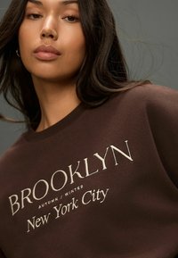 Barna pulóver, fehér hímzéssel, melyen az "BROOKLYN AUTUMN/WINTER New York City" felirat található. Sima textúrájú, kerek nyakú.
