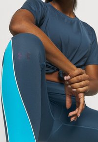 Person i blå träningstillskott, iklädd leggings med en turkos rand och Under Armour-logotyp, med händerna vilande på böjt knä.