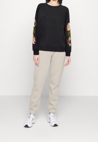 Sudadera negra con bordado floral en las mangas, combinada con pantalones de chándal beige claros. El modelo lleva zapatillas deportivas blancas.