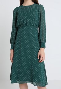 Robe verte à pois en tissu léger, avec des manches longues, une taille froncée et une jupe évasée avec des fentes latérales.