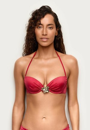 Mujer con pelo rizado que lleva un top de bikini rojo con cuello halter y un adorno de flor dorada en el centro, de pie frente a un fondo liso.