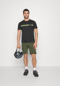 Camiseta negra con gráfico de bicicleta y franjas verdes, combinada con pantalones cortos verde oliva y sosteniendo un casco azul oscuro, zapatillas negras.