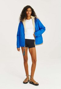 Modrá prošívaná bunda se světlým béžovým interiérem, vybavená zipem vpředu a dvěma kapsami, nošená přes bílou košili a černé kraťasy.
