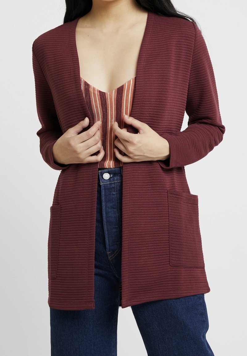 Femme portant un cardigan côtelé rouge foncé ouvert sur le devant, par-dessus un top à rayures au niveau du col et un jean bleu foncé, les mains ajustant le devant du cardigan.