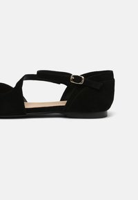 Svart mockaskor med skurna sidor, ankelrem och detaljer i guldspänne. Har en låg klack och en mjuk, slät innersula.