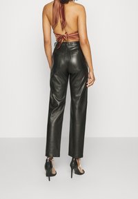 Pantaloni neri in pelle con gambe dritte, abbinati a un top in satin marrone senza schienale con un fiocco legato al collo. Le scarpe con tacco alto completano il look.