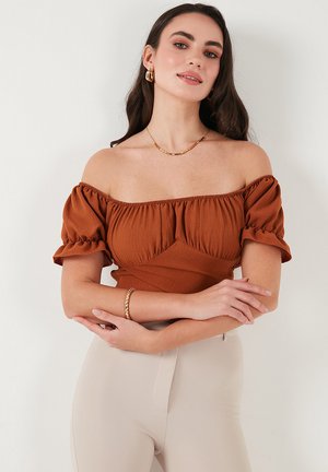 Rustrød off-shoulder top med puffærmer, tekstureret stof og ruche-detaljer, parret med højtaljede beige bukser og guldfarvede accessories.