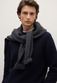 Sciarpa grigia lavorata a maglia con estremità a coste, drappeggiata casualmente intorno al collo, indossata sopra un maglione scuro e strutturato. Tessuto morbido e caldo.