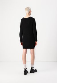 Robe noire à manches longues côtelées avec une silhouette droite, tissu texturé et fentes latérales, portée avec des chaussures noires à semelles épaisses et des chaussettes blanches.