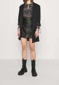 Long blazer noir, blouse noire à motifs avec nœud papillon, jupe courte noire en simili cuir avec fermeture éclair, et bottines noires épaisses.