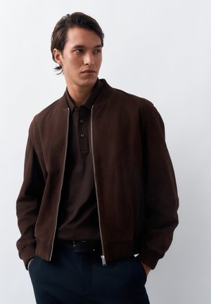 Mężczyzna ubrany w ciemnobrązową kurtkę zapinaną na zamek i koszulkę polo, z rękami w kieszeniach, patrzący na bok na jednolitym jasnym tle.