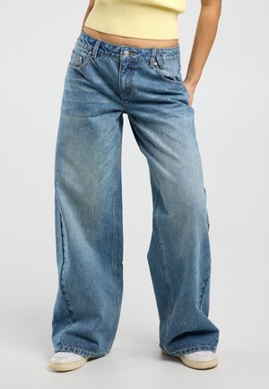 Personne portant un jean bleu en denim à jambes larges, des baskets blanches et un haut sans manches jaune, debout devant un fond blanc.