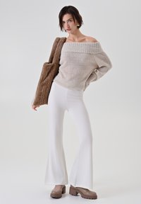 Donna che indossa un maglione beige scollato, pantaloni bianchi a zampa d'elefante, stivali marroni chunky, mentre porta a spalla una grande borsa in peluche marrone.